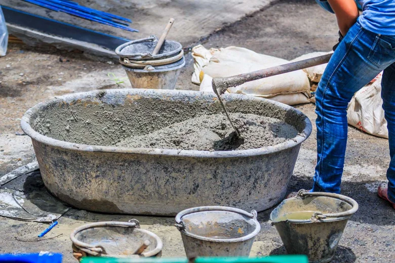 Člověk míchá beton ručně v velkém kbelíku s lopatou.