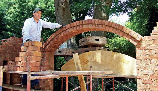 Muž staví cihlovou klenbu nad dřevěnou podložkou.