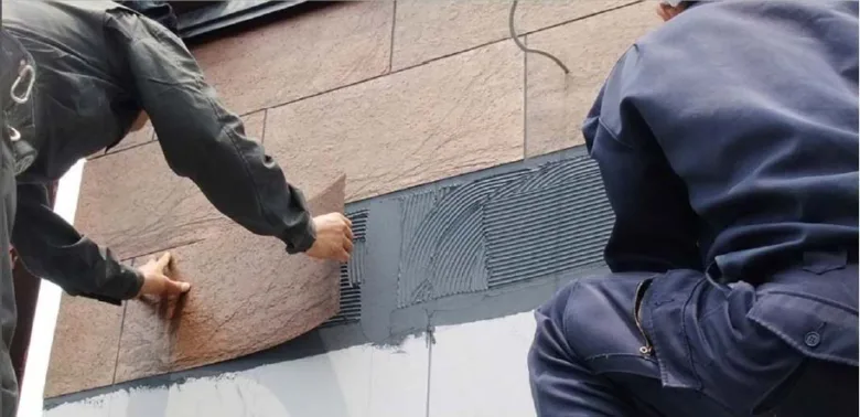 Dva dělníci montují dekorativní kámen na stěnu pomocí lepidla.