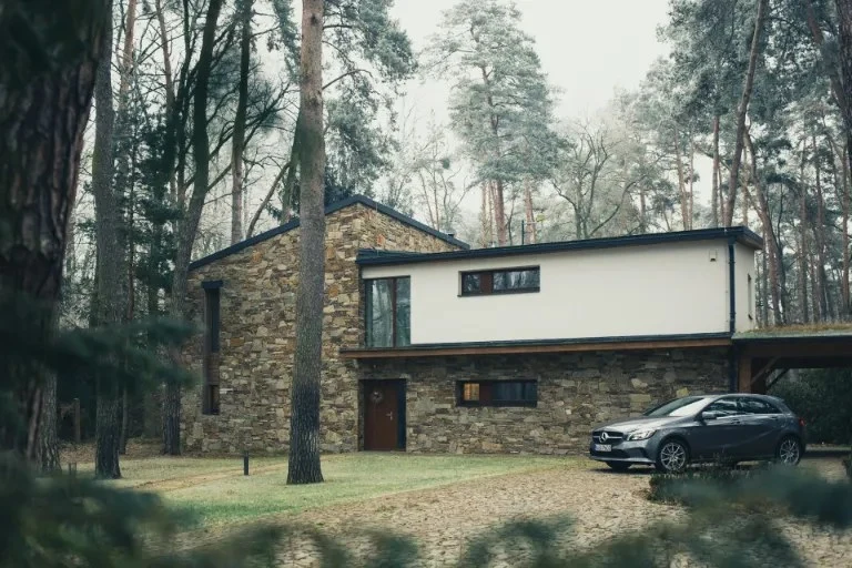 Moderní dům v lese s kamennou fasádou a parkovaným autem.