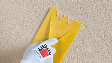 Ruka v rukavici nanáší omítku žlutou stěrnicí na zeď.