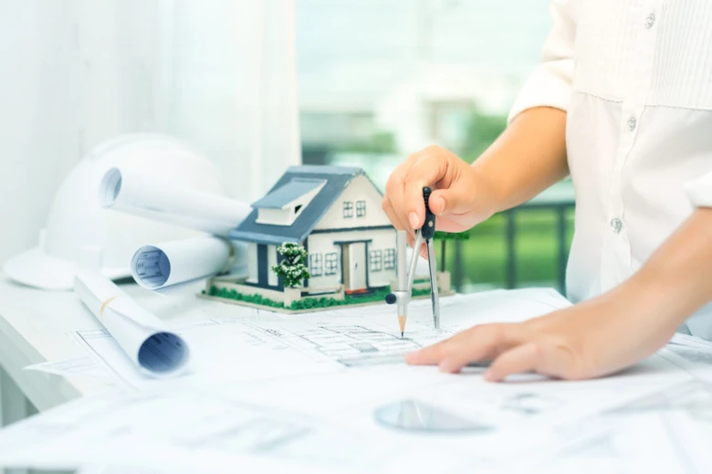 Architekt kreslí plány vedle modelu domu s kompasem.