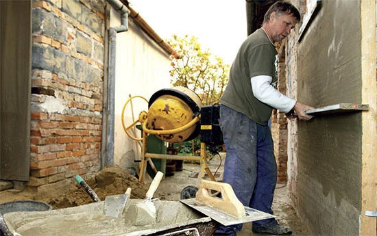 Muž nanáší omítku na zeď s použitím špachtle vedle betonového míchačky.