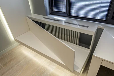 Moderní bílá skříň zakrývající topné trubky pod oknem.