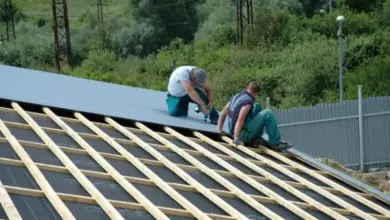 Dva dělníci upevňují vlnité plechy na dřevěné trámy střechy pomocí šroubováku.