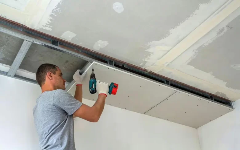 Muž montuje sádrokartonový strop pomocí vrtáku na kovovém rámu.