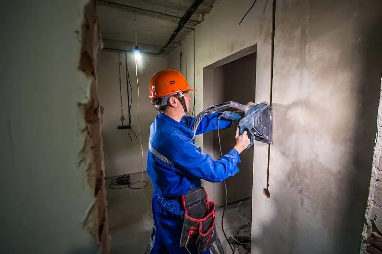 Stavebník v modrém obleku děrá stěnu pro instalaci kabeláže.