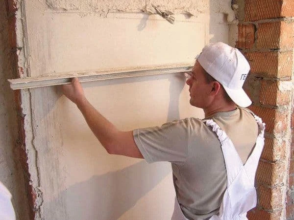 Stavebník nanáší omítku na stěnu pomocí pravítka pro vyrovnání povrchu.