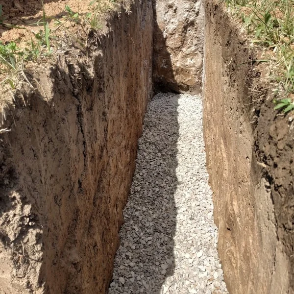 Výkop s kamenivem pro instalaci drenážního potrubí kolem domu.