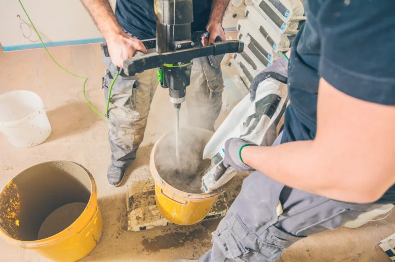 Dva dělníci míchají beton v žlutém kbelíku pomocí vrtačky.