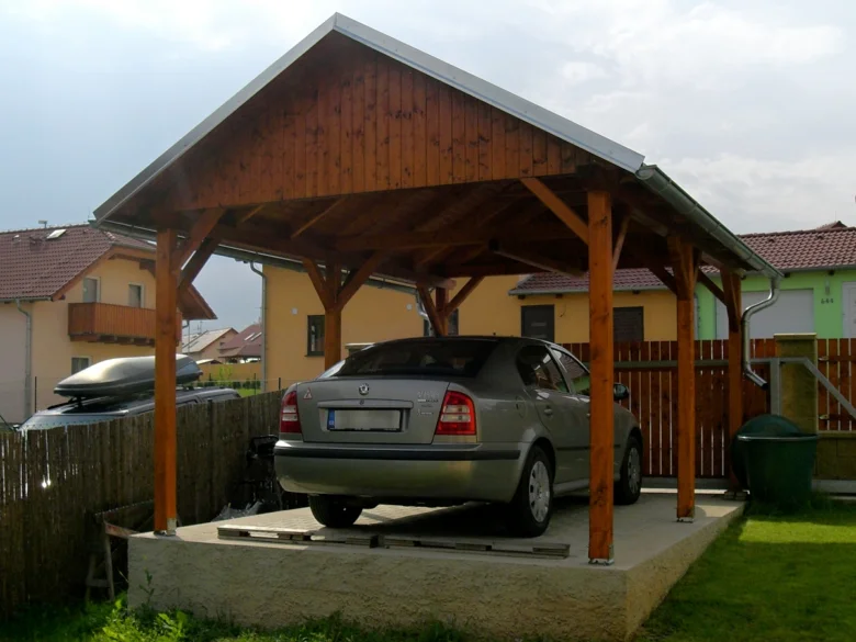 Hotový dřevěný přístřešek pro auto s stříbrným autem pod ním na betonovém základu v rezidenční oblasti.