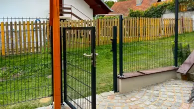 Ocelový plot s bránou vedle dřevěného plotu v zahradním pozadí.