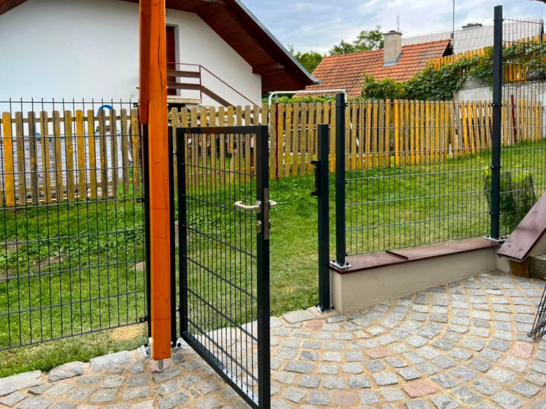 Ocelový plot s bránou vedle dřevěného plotu v zahradním pozadí.