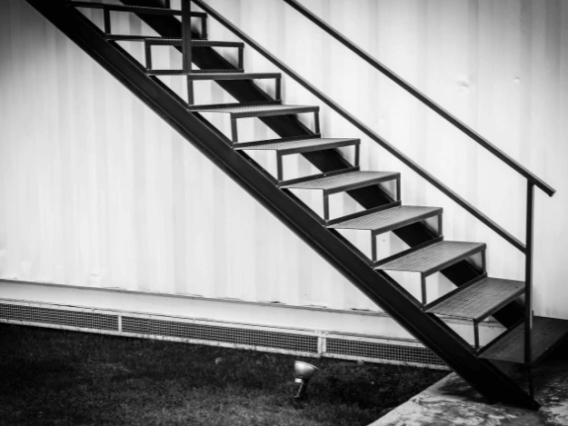 Černobílá fotografie kovového venkovního schodiště s mřížovými stupni.