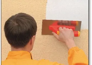 Muž aplikuje válcovou omítku na fasádu domu pomocí valčkového nástroje.