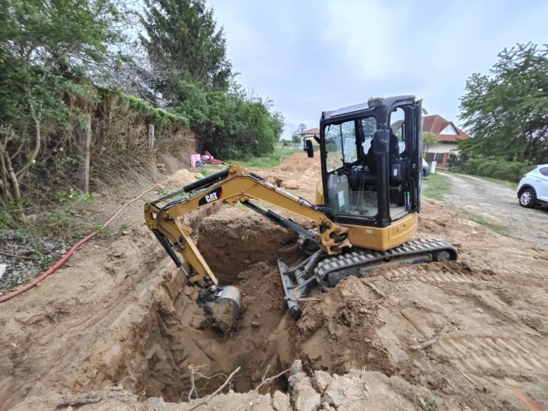 Žlutý bagr vykopává díru pro venkovní záchod v zahradě.