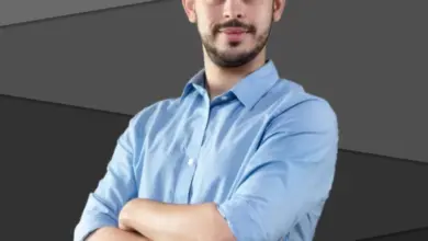 Muž v modré košili s kříženýma rukama na tmavém pozadí.