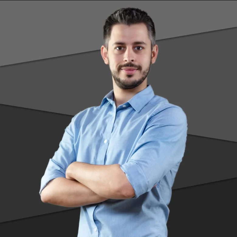 Muž v modré košili s kříženýma rukama na tmavém pozadí.