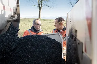 Dva pracovníci v oranžových vestách u asfaltové drti venku.