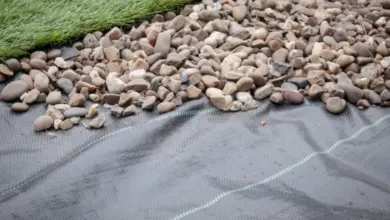 Černá geotextilní fólie pod drženým kamenem s trávníkem v pozadí.