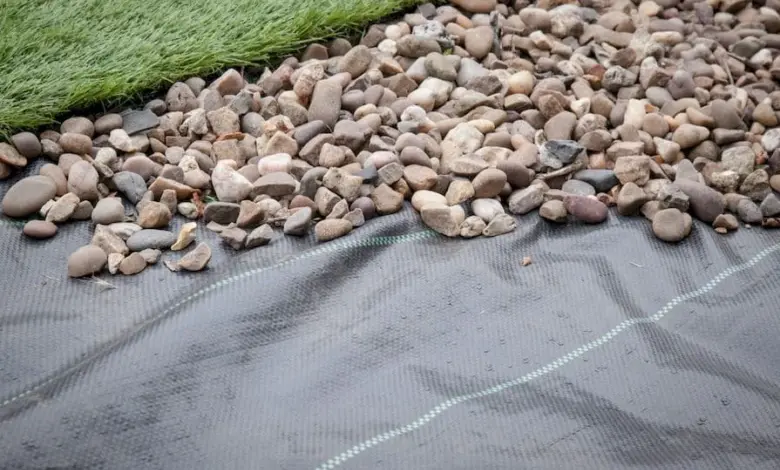 Černá geotextilní fólie pod drženým kamenem s trávníkem v pozadí.