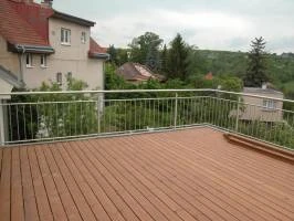 Moderní kovové zábradlí na dřevěném balkóně s výhledem do zelené krajiny.