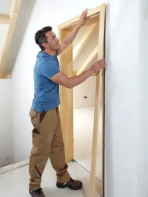 Muž montuje dřevěný rámeček interiérových dveří do stěny.