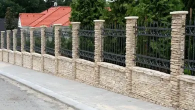 Kamenný plot s kovovou konstrukcí na pozadí domů.