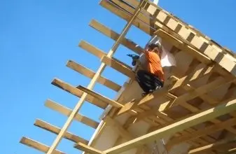 Stavebník montuje trámy sedlové střechy na dřevěném nosníku.