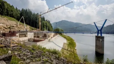 Stavba betonové hráz u jezera s kranem v lesnatém prostředí.