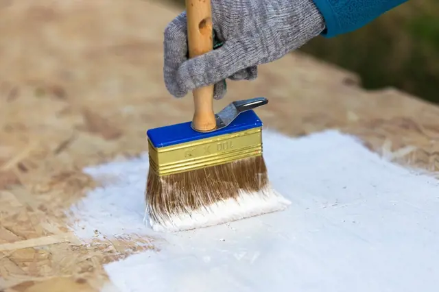 Ruka v rukavici natírá OSB desku bílou barvou štětcem.
