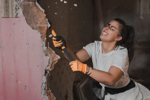 Žena bourá nosnou stěnu v budově odhalující cihlovou konstrukci.