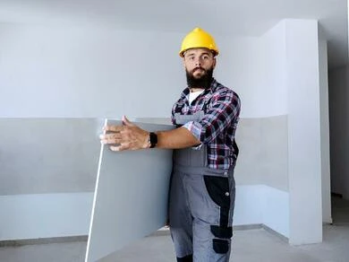 Stavebník drží sádrokartonovou desku v prázdné místnosti.
