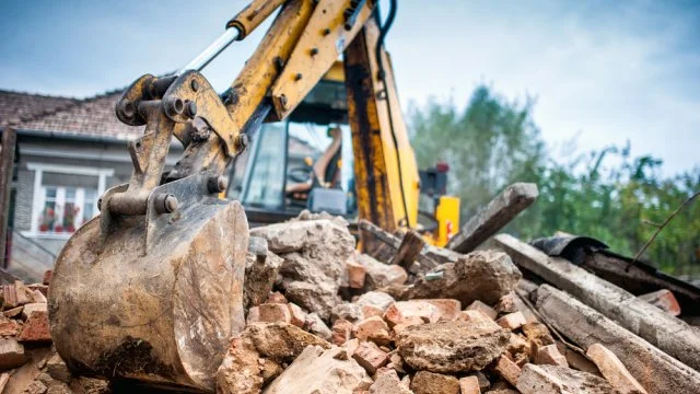 Žlutý bagr rozebírá trosku zbourané kůlny na staveništi.