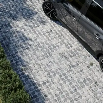 Šedá auto stojí na dlažbě z kamenných kostek vedle keřů.