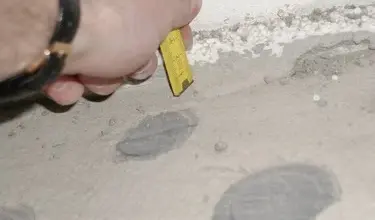 Ruka měří tloušťku betonu žlutým pravítkem na hrubé podlaze.