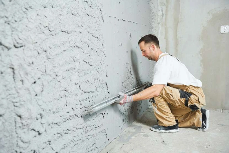 Muž v kombinéze hladí omítku na vlhkém zdi ve sklepě.