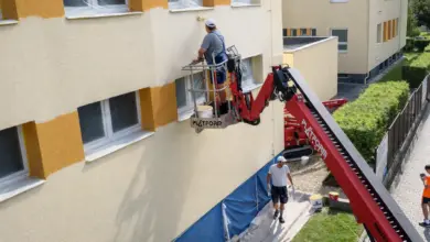 Muž maluje fasádu domu pomocí zdviha na slunci.