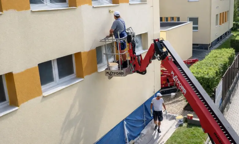 Muž maluje fasádu domu pomocí zdviha na slunci.