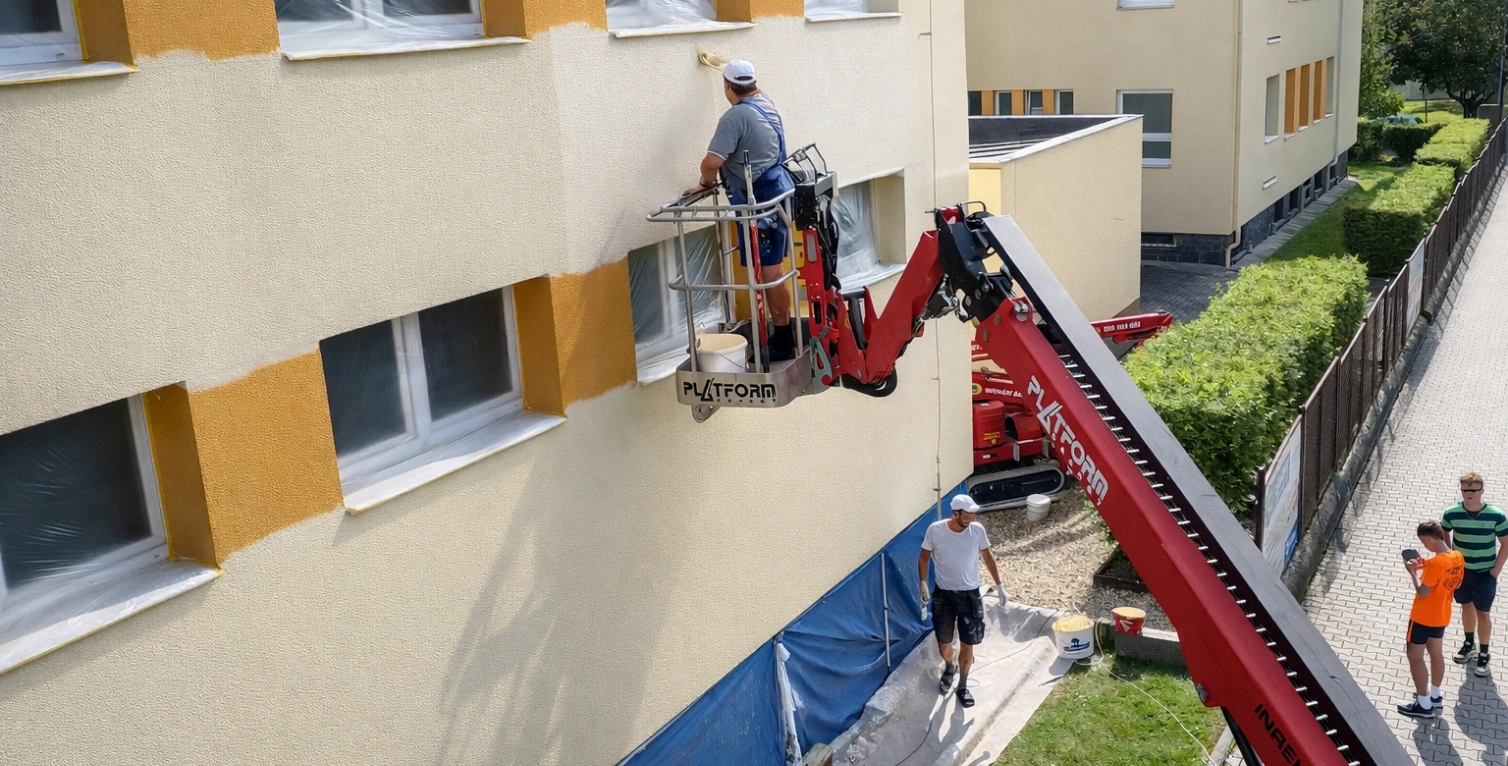Muž maluje fasádu domu pomocí zdviha na slunci.