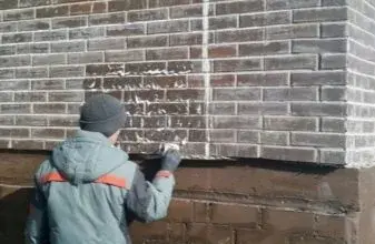 Muž čistí bílé výkvěty z cihlové zdi pomocí škrabky.
