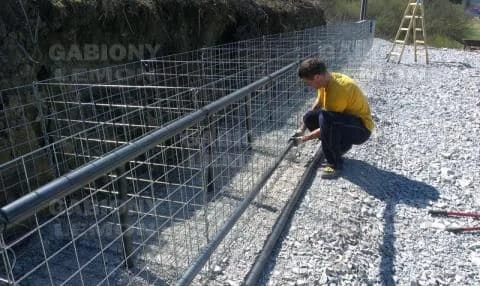 Muž montuje ocelový plot z kbelíků s kamením na skále.