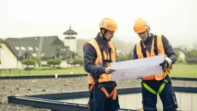Dva stavebníci na ploché střechě studují plány pro instalaci vpustí.